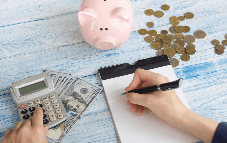 Person planning their budget on a notepad, with a calculator, cash, and piggy bank nearby